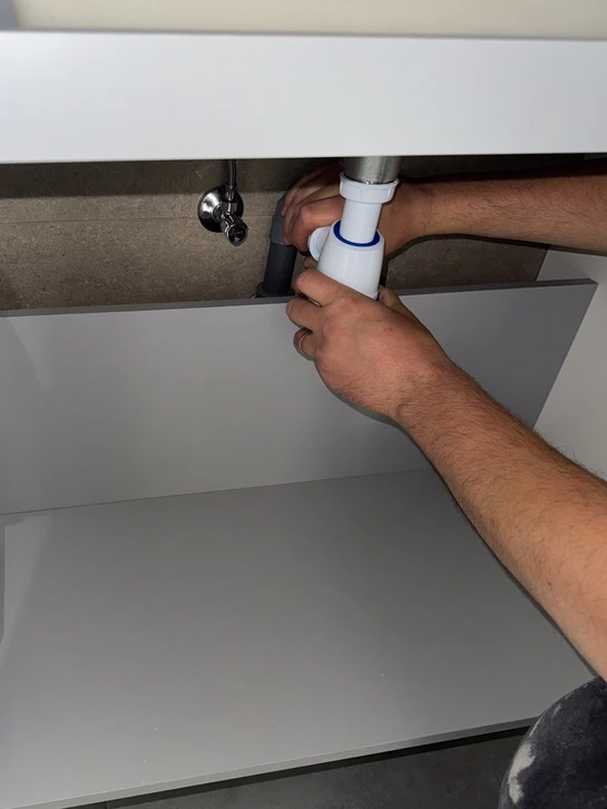 Photographie d'un plombier-chauffagiste dans une salle de bains en cours de rénovation, expliquant les détails du projet à un client. L'artisan pointe des éléments du chantier (canalisation, installation de douche, équipements sanitaires) tandis que le client l'écoute attentivement. L'environnement montre un chantier organisé et contrôlé, avec des outils professionnels visibles. La scène doit transmettre la confiance, la clarté des explications et la maîtrise professionnelle du projet.